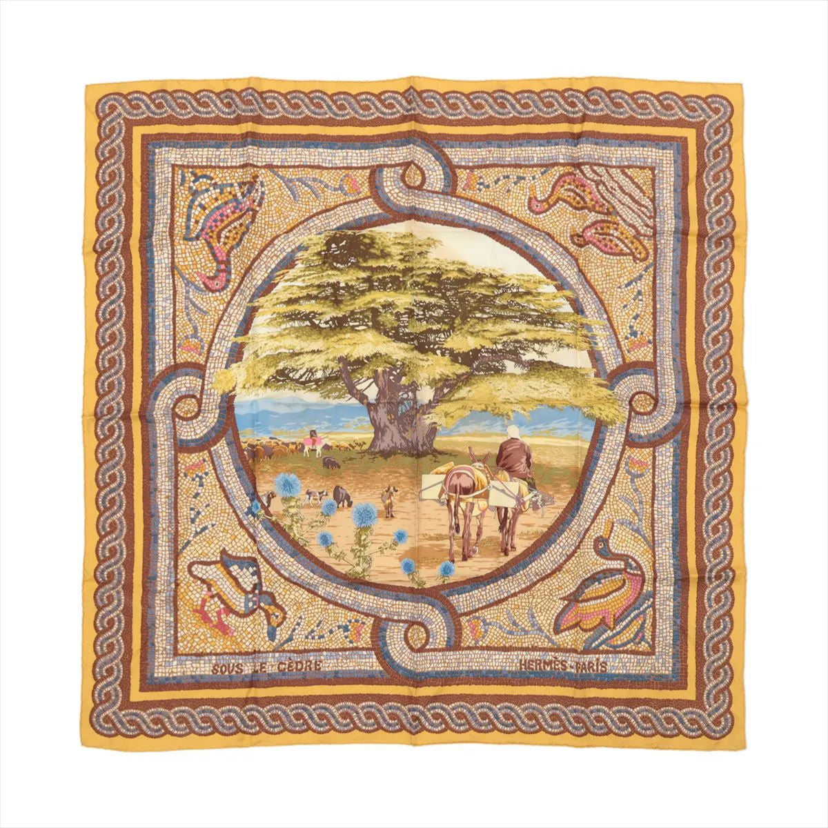 Flat-lay image of Hermès Carré 90 "Sous le Cèdre" silk scarf featuring a large cedar tree and pastoral figures in the center, with a brown, gold, and multicolor mosaic border design.