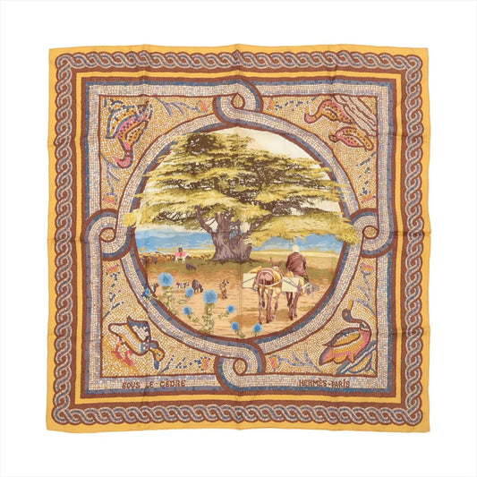 Flat-lay image of Hermès Carré 90 "Sous le Cèdre" silk scarf featuring a large cedar tree and pastoral figures in the center, with a brown, gold, and multicolor mosaic border design.