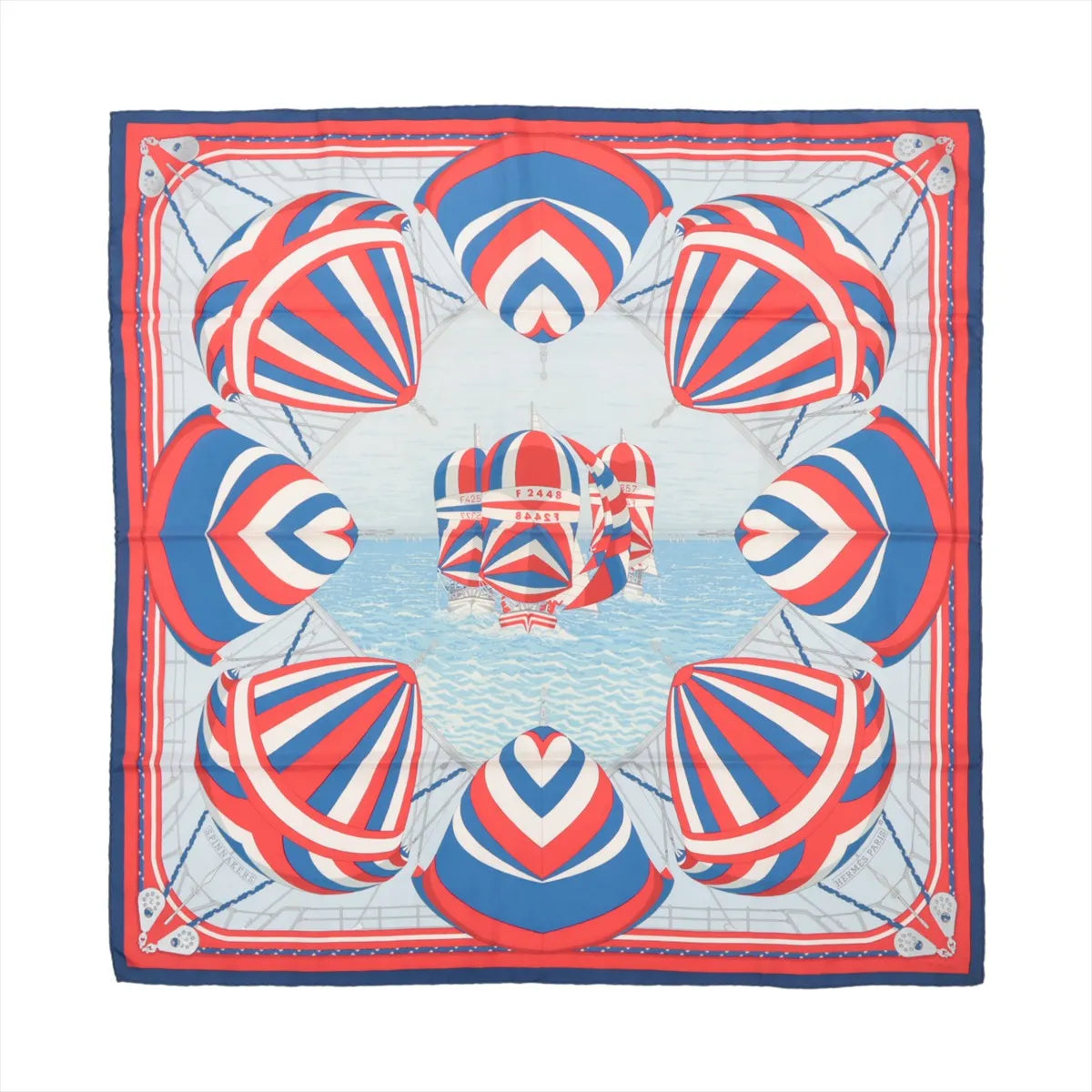 Flat-lay of a Hermès Carré 90 "Spinnakers" silk scarf featuring a vibrant nautical design with red, white, and blue striped spinnaker sails over a sea-blue background, bordered in coordinating colors.