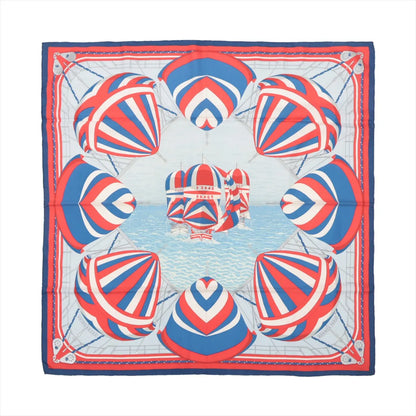 Flat-lay of a Hermès Carré 90 "Spinnakers" silk scarf featuring a vibrant nautical design with red, white, and blue striped spinnaker sails over a sea-blue background, bordered in coordinating colors.