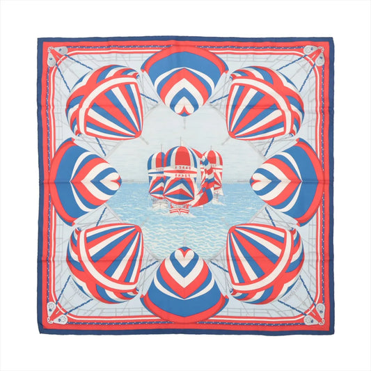 Flat-lay of a Hermès Carré 90 "Spinnakers" silk scarf featuring a vibrant nautical design with red, white, and blue striped spinnaker sails over a sea-blue background, bordered in coordinating colors.