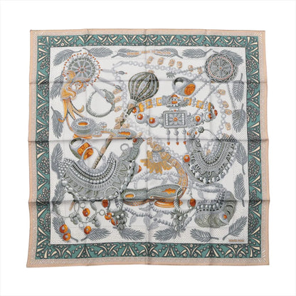 Hermès Carré 90 "Zenobie Reine de Palmyre" silk scarf in beige and green featuring intricately illustrated ancient jewelry in silver, gold, and orange tones, framed by a patterned green and beige border with palm fronds and decorative motifs.