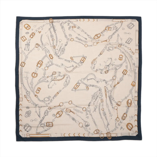 Flat-lay of a Hermès Carré Géant "Malines Chevaux Déchène" scarf in beige with navy blue border, featuring detailed line drawings of horses formed by intertwined bridles and tack in gray and gold tones, printed on a soft cashmere and silk blend.