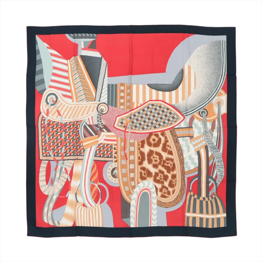 Hermès Carré Géant "La Selle Imaginaire" scarf in red, made from a cashmere and silk blend, showcasing a colorful abstract saddle-themed design with patterns in orange, beige, blue, and gray, bordered by a solid dark navy edge, displayed flat against a white background.