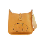 Camel brown pebbled leather Hermès Evelyne GM shoulder bag with perforated H logo and beige canvas strap on white background.