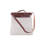 Hermès Herbag GM beige canvas bag with brown leather flap, top handle, long shoulder strap and gold-tone circular closure.
