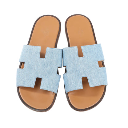 Hermès Izmir sandals in light blue denim featuring wide straps with the signature “H” cut-out design, tan leather footbeds, and dark brown soles. Top view, size 43 EU.