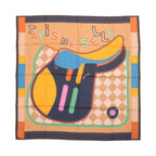Hermès Carré 90 double-face silk scarf laid flat with large multicolor saddle illustration, “Tapis de Selle” text, and orange border on a beige patterned background.