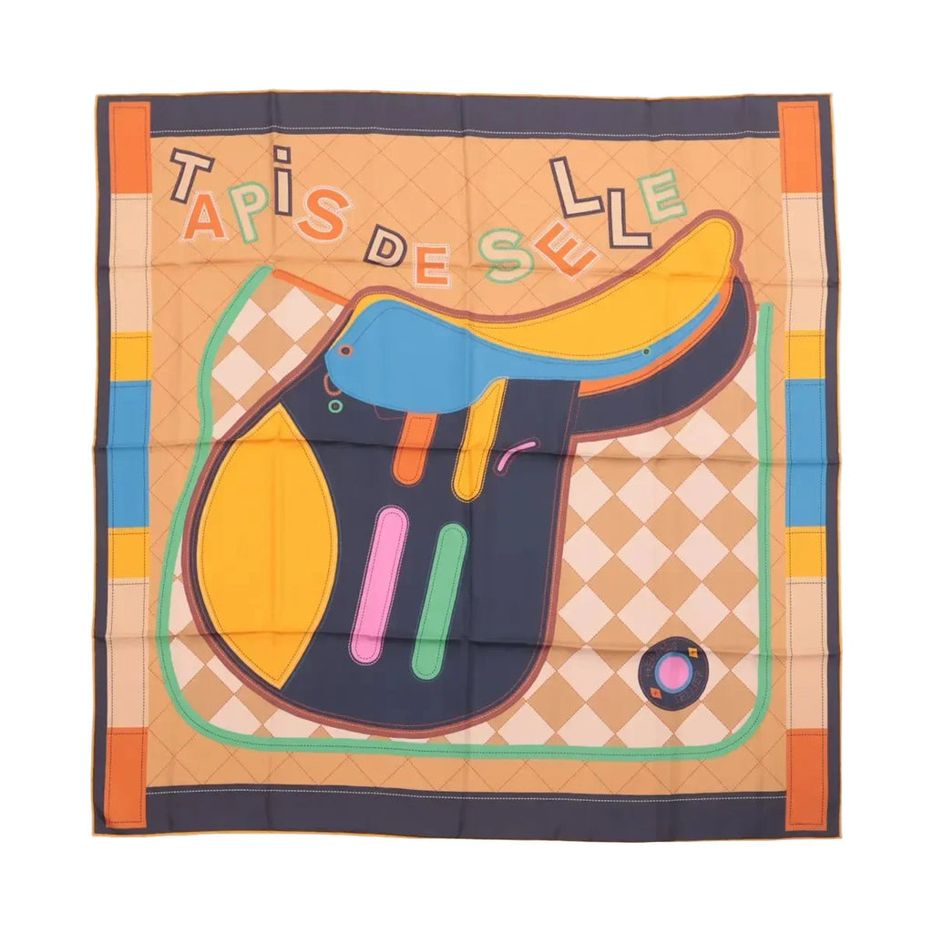 Hermès Carré 90 double-face silk scarf laid flat with large multicolor saddle illustration, “Tapis de Selle” text, and orange border on a beige patterned background.