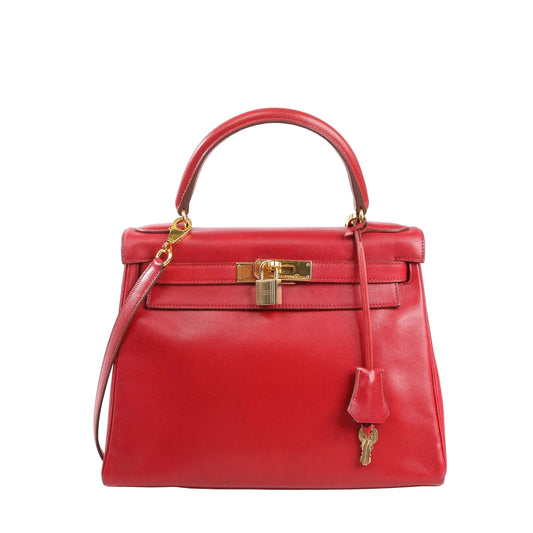 Front view of a red Hermès Kelly 28 Box leather handbag with top handle, detachable shoulder strap, gold turn-lock hardware, and hanging clochette with keys on a white background.