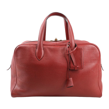 Front view of a Hermès Victoria 43 travel bag in Rouge H Taurillon Clemence leather, featuring dual rounded top handles, gold-tone hardware, leather clochette, and soft pebble-grain texture.