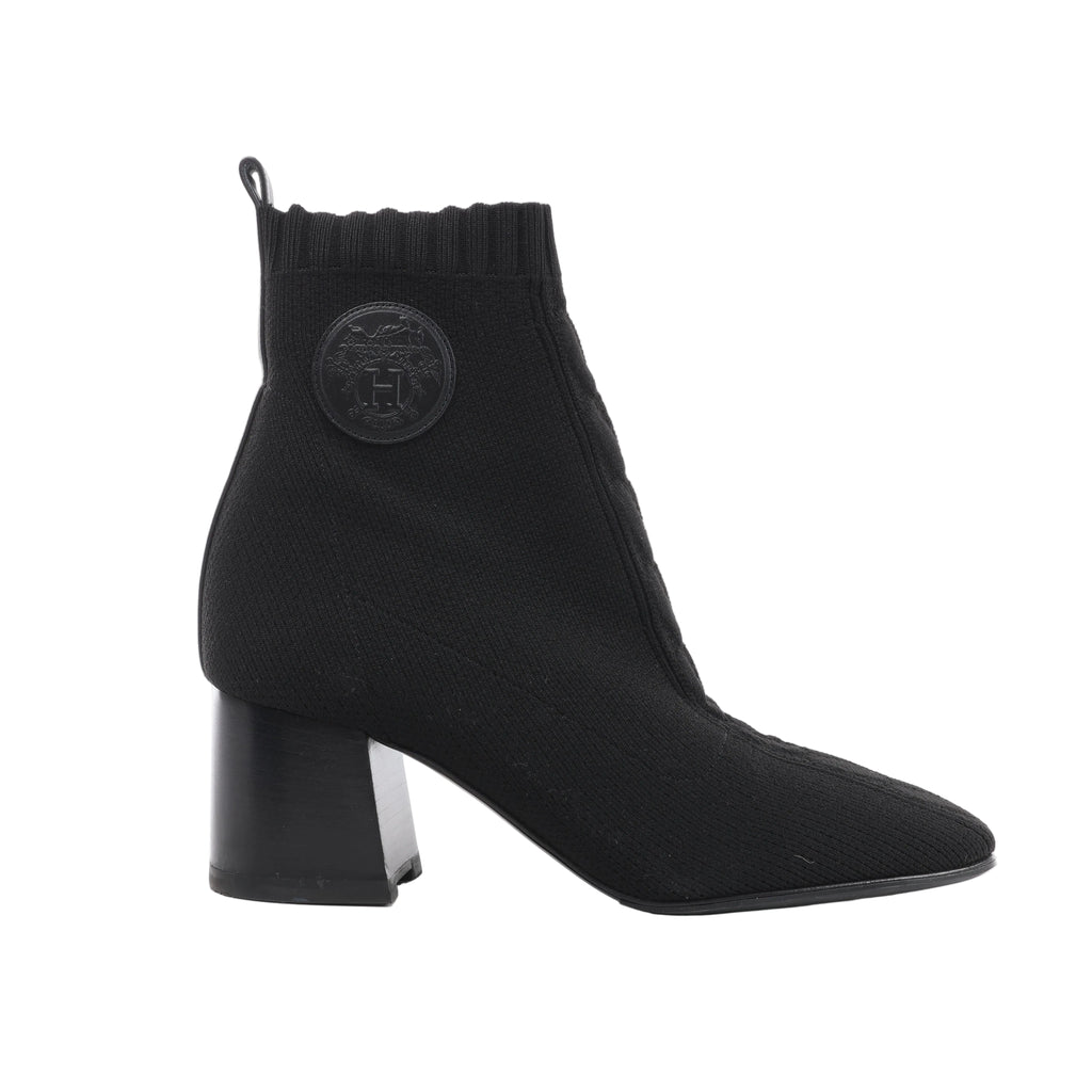 Side view of a black Hermès Volver knit ankle boot with a ribbed cuff, round tonal leather emblem, rear pull tab and block heel.