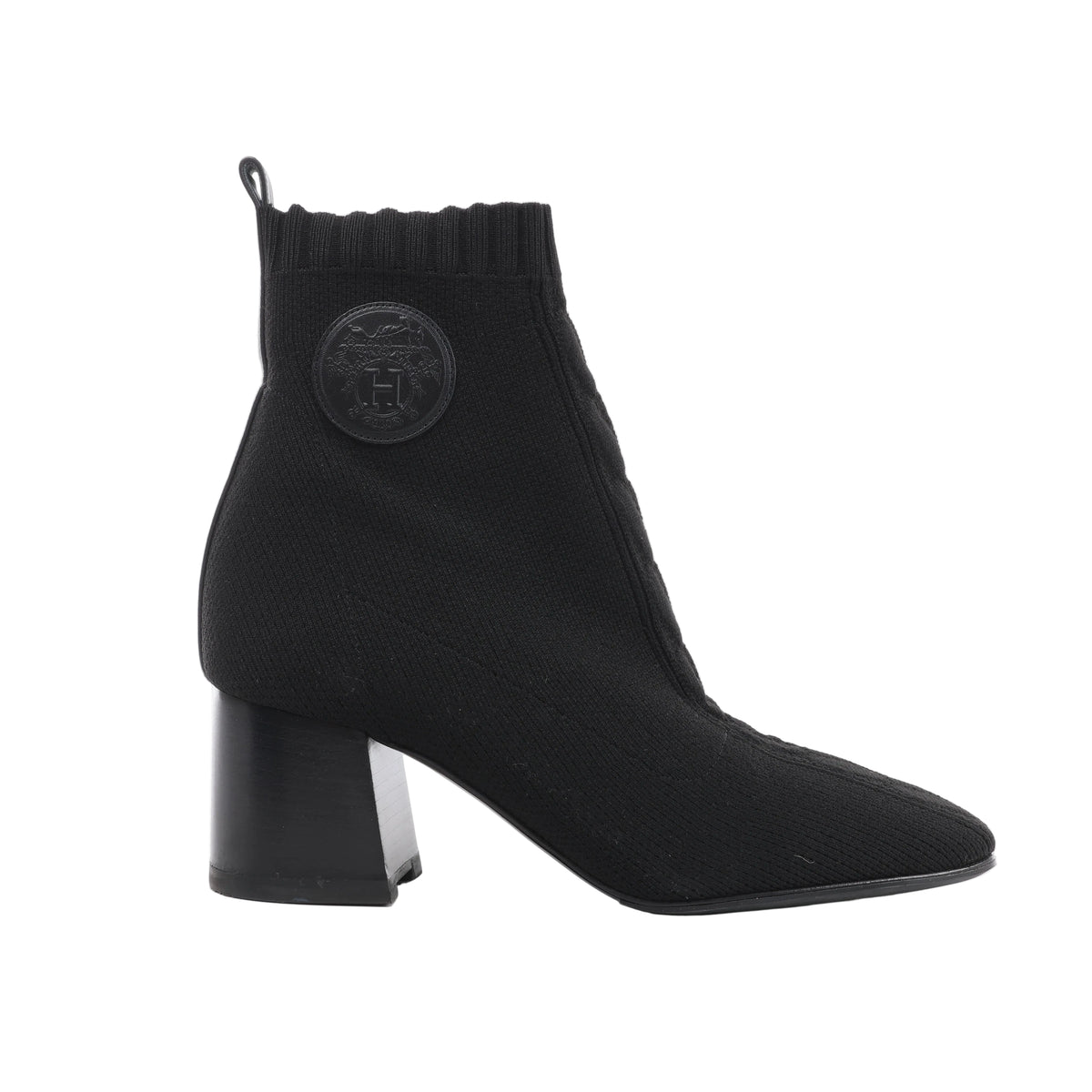 Side view of a black Hermès Volver knit ankle boot with a ribbed cuff, round tonal leather emblem, rear pull tab and block heel.