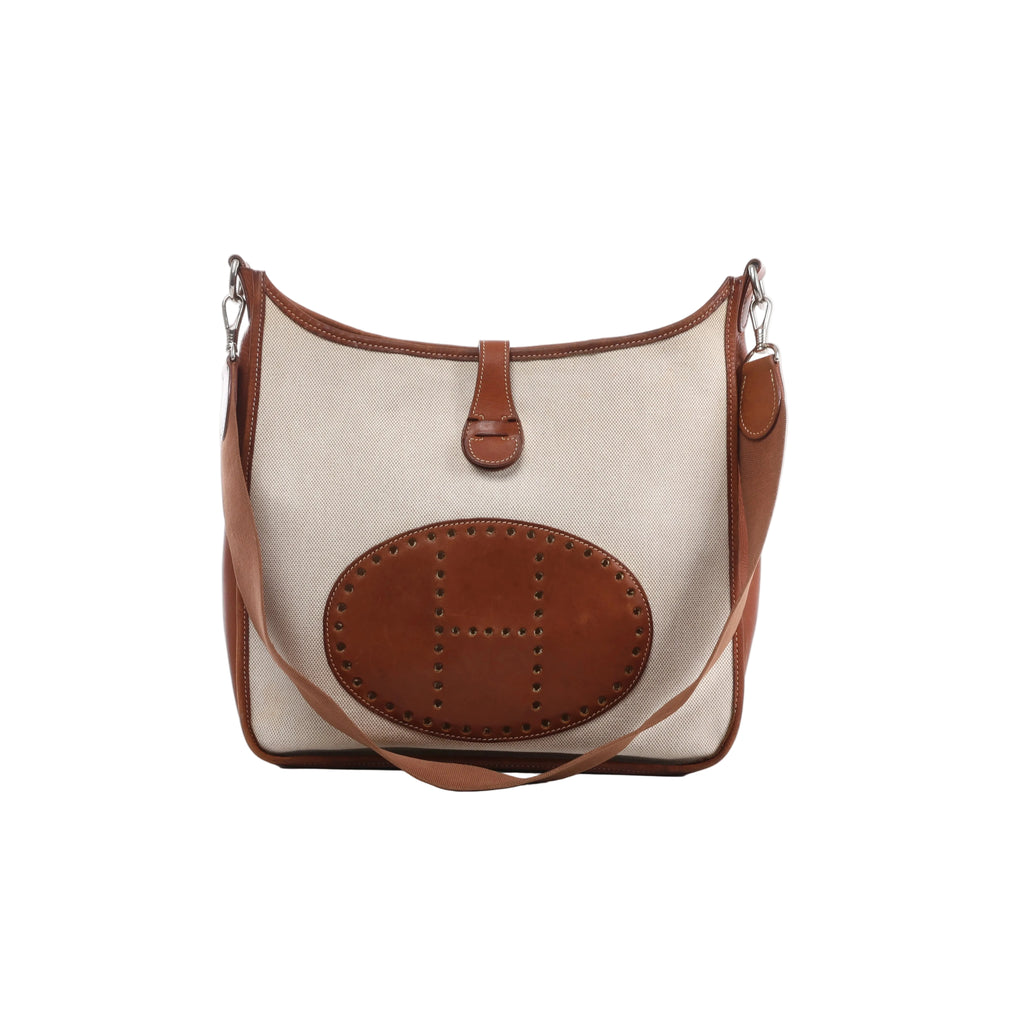 Front view of Hermès Evelyne I bag in beige canvas with brown leather trim, oval perforated H panel, brown shoulder strap and silver-tone hardware.