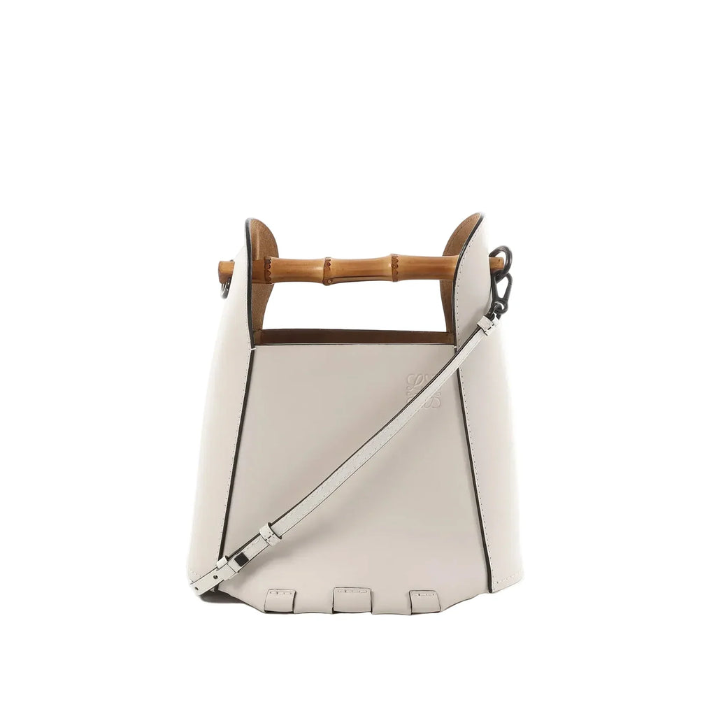 White leather LOEWE bucket handbag with bamboo top handle, detachable shoulder strap, black edge trim, and embossed logo on front.