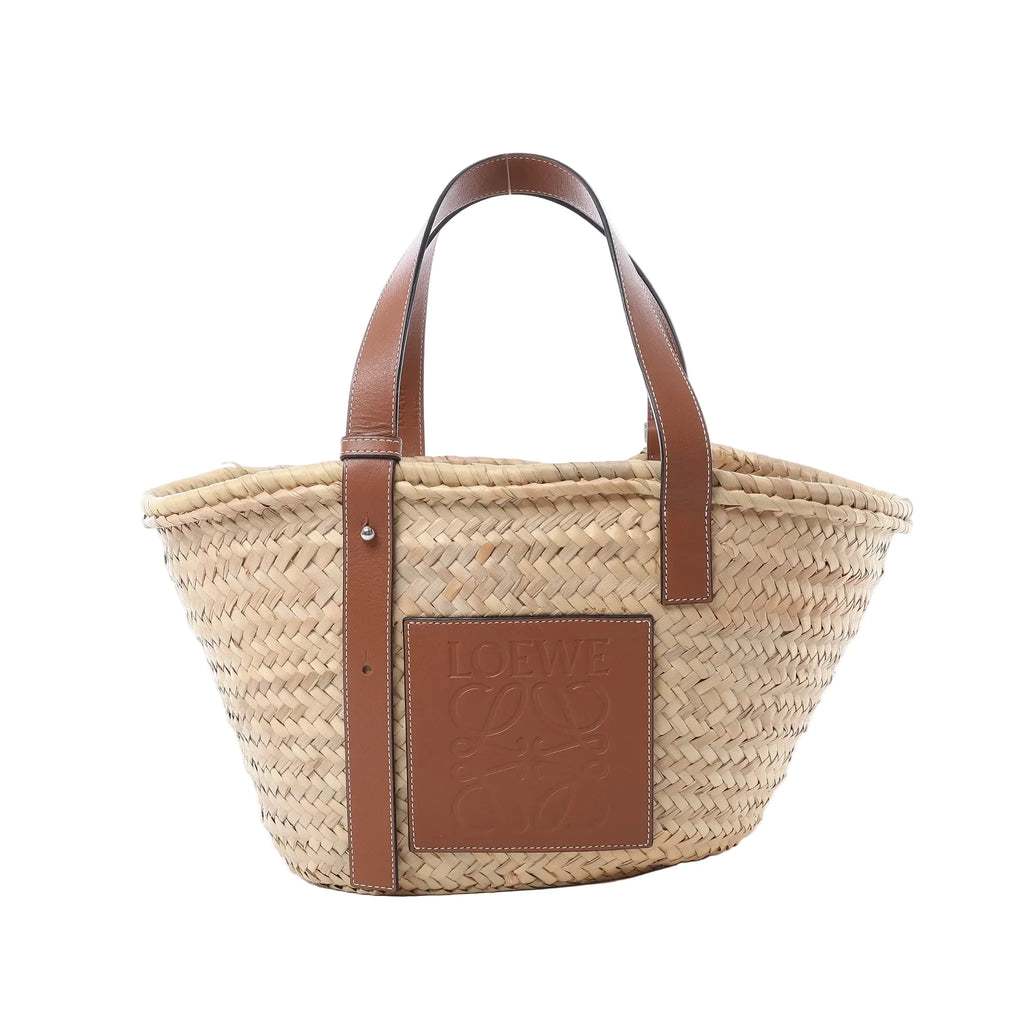 LOEWE Basket Medium handbag in beige woven raffia with brown leather shoulder straps, embossed leather logo patch, and open-top tote shape on white background.