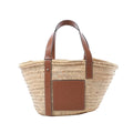 LOEWE Basket Medium handbag in beige woven raffia with brown leather shoulder straps, embossed leather logo patch, and open-top tote shape on white background.