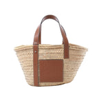 LOEWE Basket Medium handbag in beige woven raffia with brown leather shoulder straps, embossed leather logo patch, and open-top tote shape on white background.