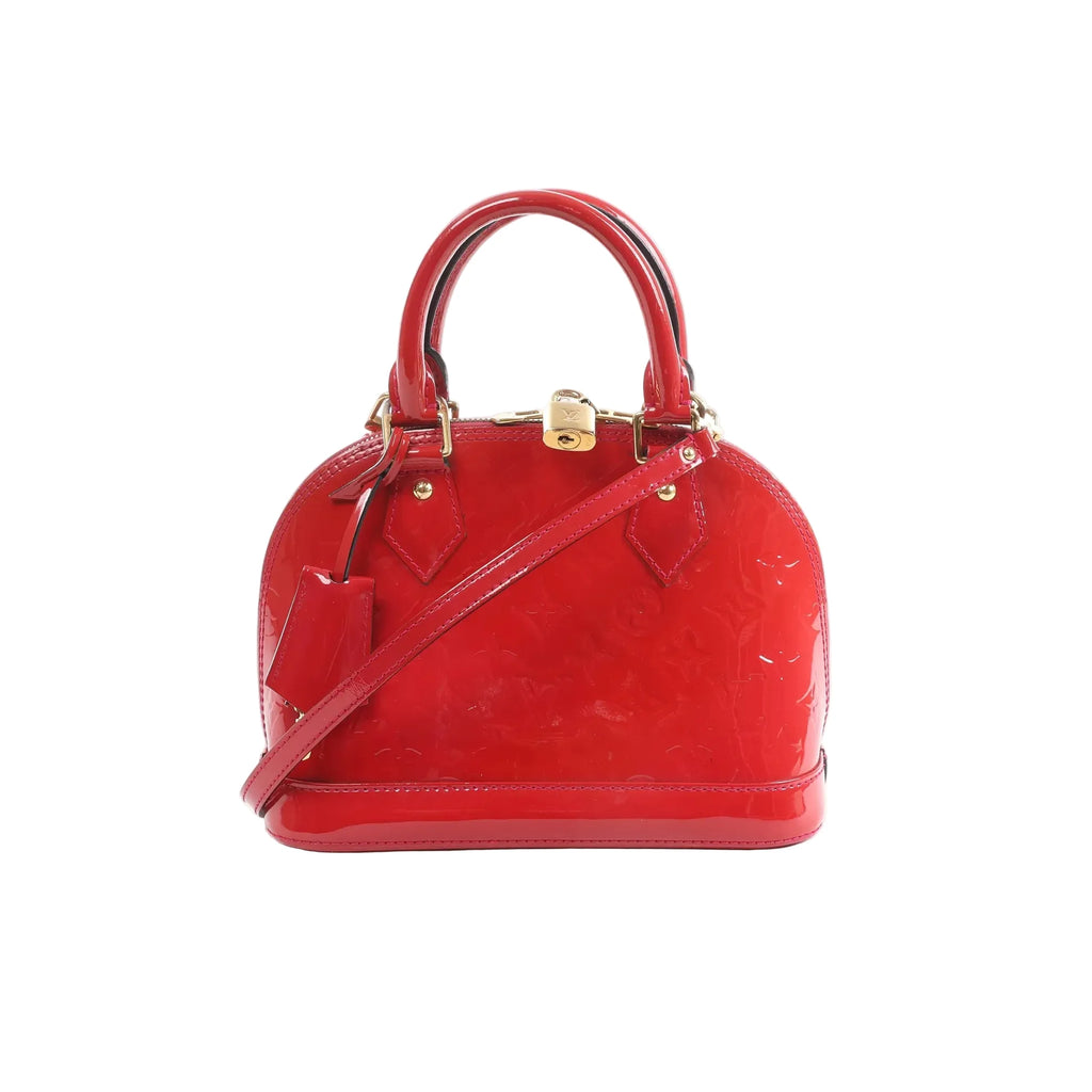 Red glossy Louis Vuitton Alma BB Monogram Vernis handbag with top handles, detachable shoulder strap, gold-tone padlock, and hanging luggage tag on a white background.