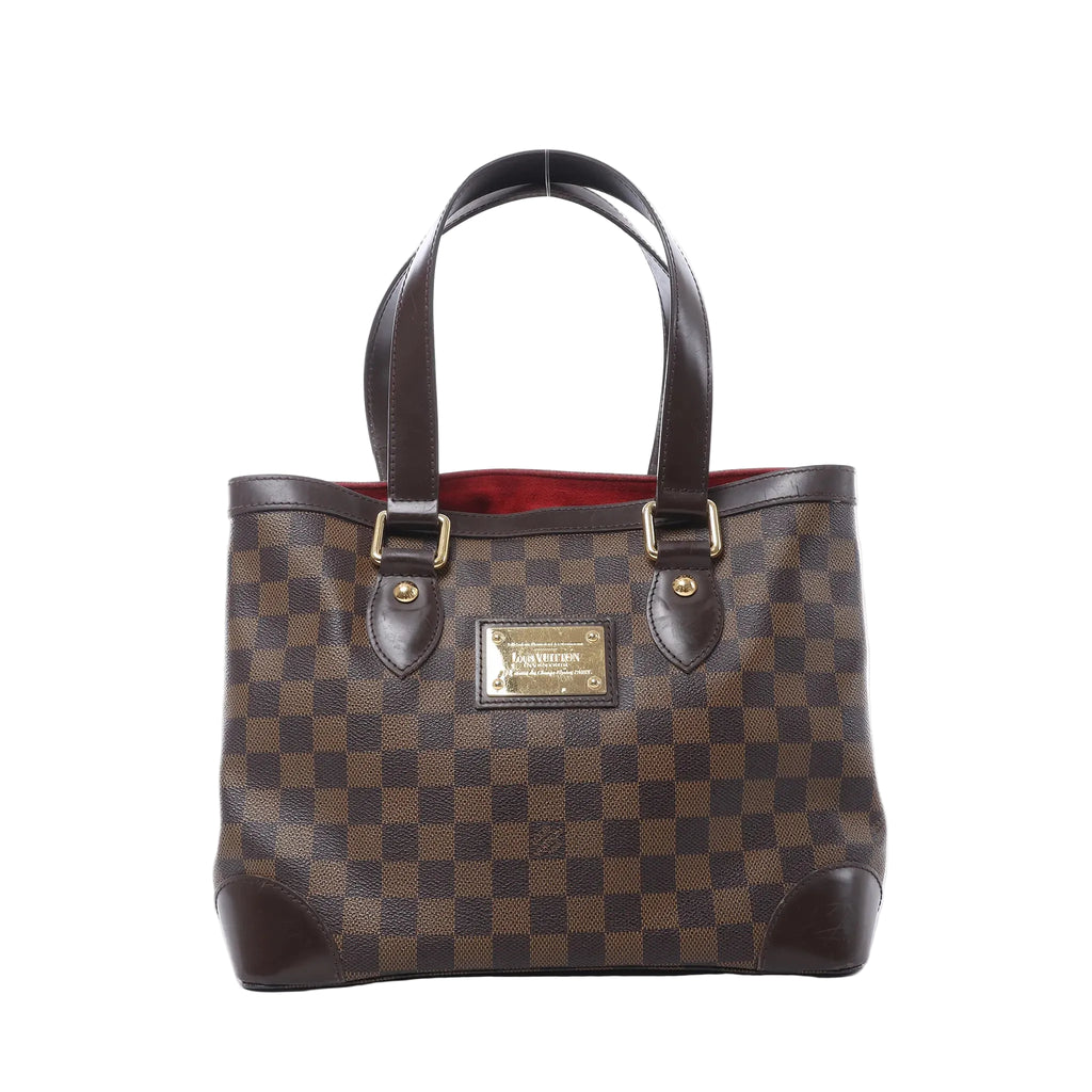 Louis Vuitton Hampstead PM shoulder bag in brown Damier Ebene canvas with dark leather handles, gold-tone hardware, front logo plaque, and red interior visible on white background.
