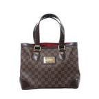 Louis Vuitton Hampstead PM shoulder bag in brown Damier Ebene canvas with dark leather handles, gold-tone hardware, front logo plaque, and red interior visible on white background.