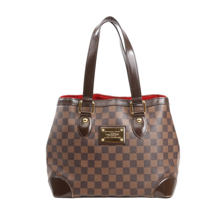 Louis Vuitton Hampstead PM shoulder bag in brown Damier Ebene canvas with dual dark brown leather straps, gold-tone plaque, and red interior lining, displayed on a white background.