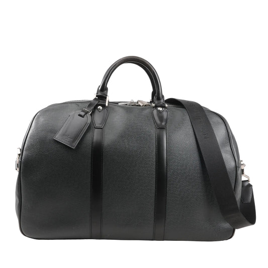 Front view of a Louis Vuitton Kendall PM M30122 travel bag in black Taiga leather featuring structured dual top handles, a detachable black shoulder strap, silver-tone hardware, and matching leather luggage tag.
