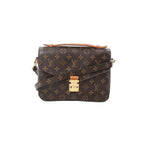 Front view of Louis Vuitton Monogram canvas Pochette Métis MM with tan leather top handle, gold-tone S-lock clasp, and crossbody strap on white background.