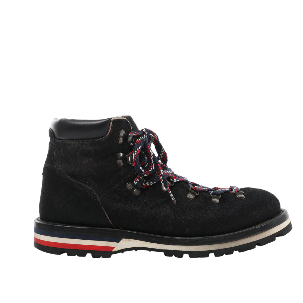 Side view of a black Moncler ankle boot with a hair-textured upper, metal lace hooks, tricolour laces and a lugged sole with striped heel detailing.