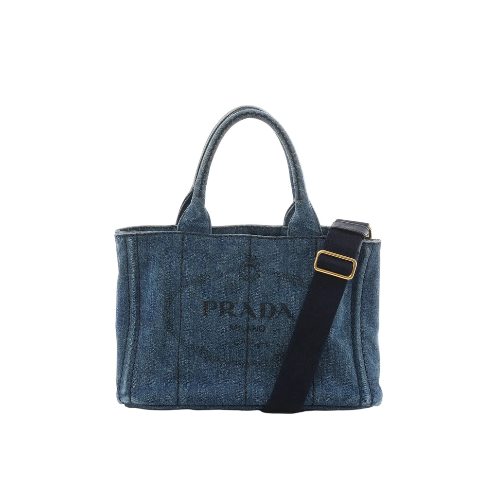 Blue denim Prada Canapa tote handbag with top handles, black adjustable shoulder strap with gold buckle, contrast stitching, and PRADA Milano logo on front.