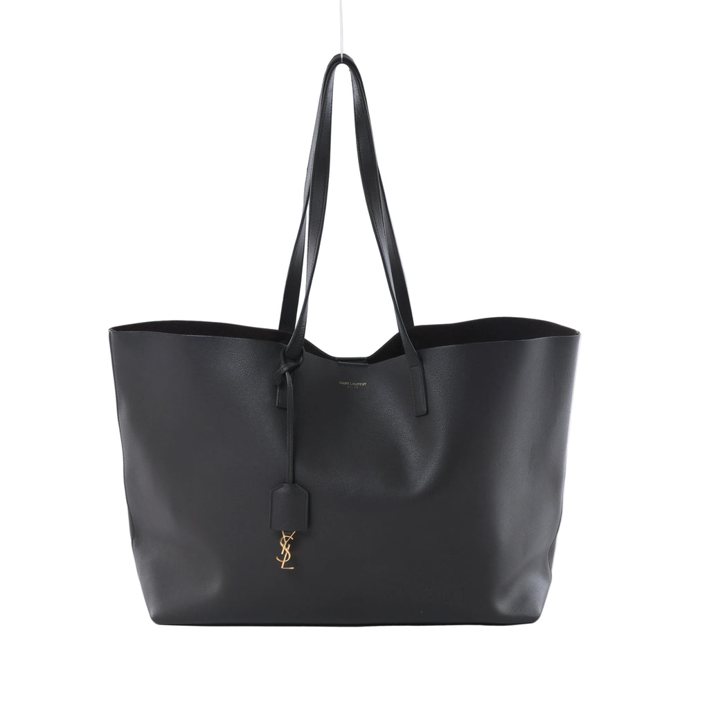 Gray Saint Laurent Sac Shopping leather tote with black shoulder straps and a gold-tone YSL charm, shown from the front on a light background.