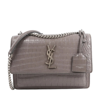 Front view of the Saint Laurent Paris Sunset Medium Mock Croc Shoulder Bag in gray, showcasing embossed crocodile-textured leather, silver-tone YSL logo hardware, chain shoulder strap, and leather tag detail, set against a white background.