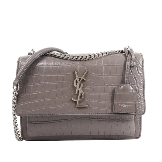 Front view of the Saint Laurent Paris Sunset Medium Mock Croc Shoulder Bag in gray, showcasing embossed crocodile-textured leather, silver-tone YSL logo hardware, chain shoulder strap, and leather tag detail, set against a white background.