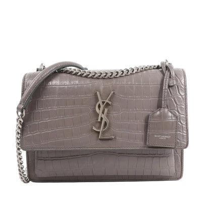 Front view of the Saint Laurent Paris Sunset Medium Mock Croc Shoulder Bag in gray, showcasing embossed crocodile-textured leather, silver-tone YSL logo hardware, chain shoulder strap, and leather tag detail, set against a white background.