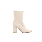 Side view of a Valentino Garavani ivory leather boot with a pointed toe, tall block heel and a row of metallic pyramid studs along the back.