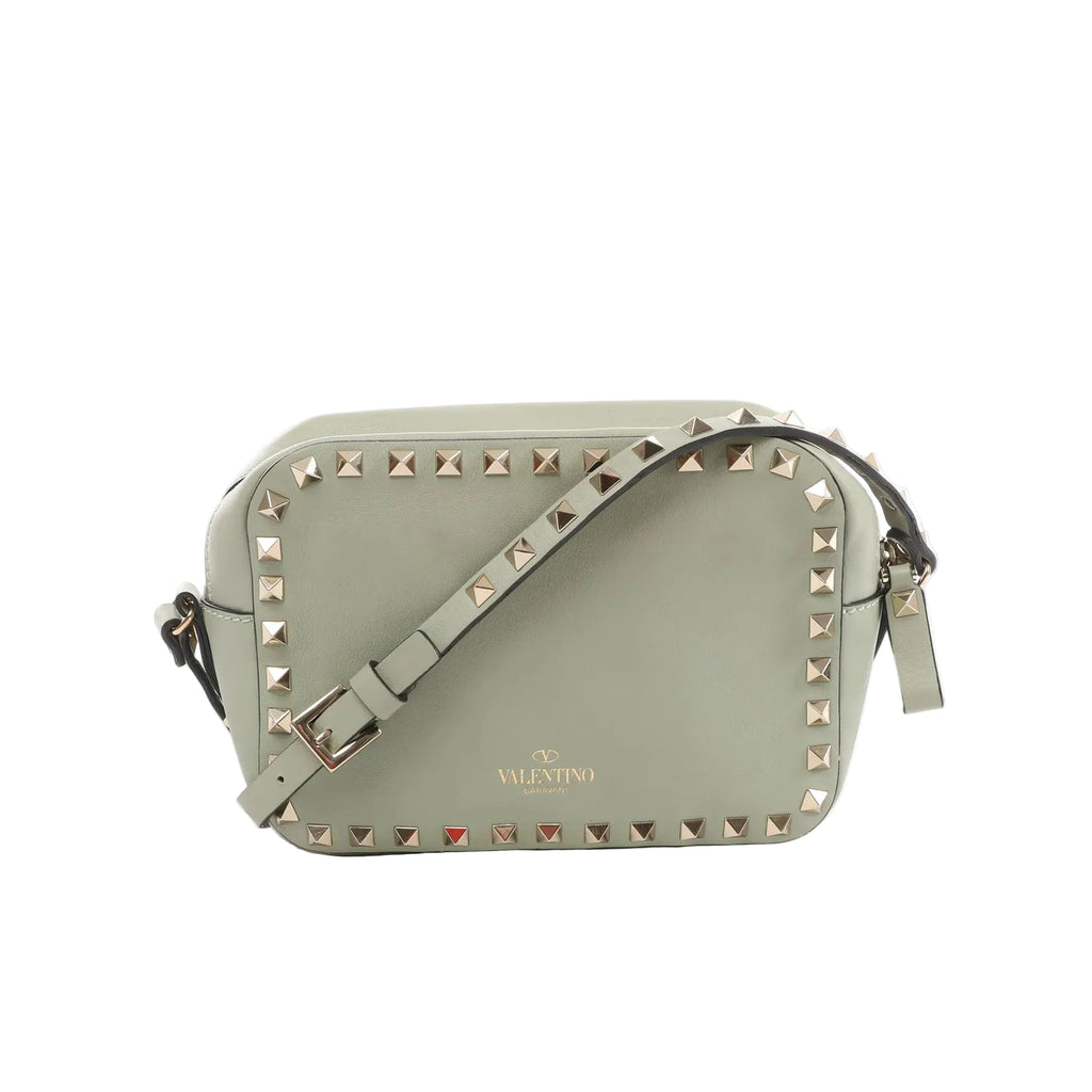 Sage green leather Valentino Garavani crossbody bag with gold-tone pyramid studs around the edge, adjustable strap, and zip-top closure on a white background.