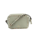 Sage green leather Valentino Garavani crossbody bag with gold-tone pyramid studs around the edge, adjustable strap, and zip-top closure on a white background.