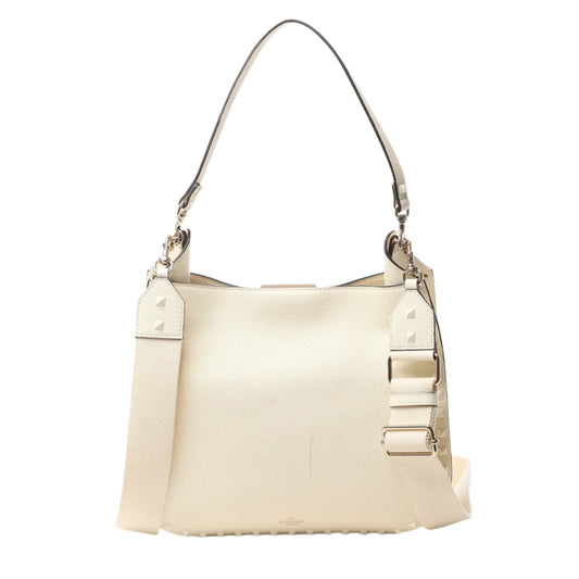 Front view of Valentino Garavani Rockstud Leather Shoulder Bag in ecru grained leather with tonal pyramid stud trim, top handle, and wide adjustable shoulder strap, displayed against a white background.