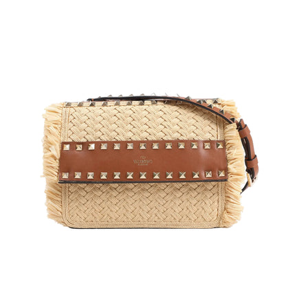 Valentino Garavani shoulder bag crafted from beige woven straw with brown leather flap and strap, adorned with signature gold-tone Rockstuds and fringe trim along the sides, displayed against a white background.