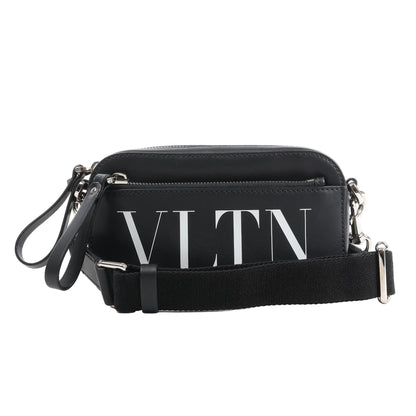 Front view of the Valentino Garavani VLTN Leather Shoulder Bag in black, featuring smooth leather exterior, bold white VLTN logo print, front zip pocket, detachable black fabric strap, and silver-tone hardware, displayed against a white background.
