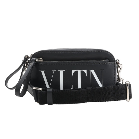 Front view of the Valentino Garavani VLTN Leather Shoulder Bag in black, featuring smooth leather exterior, bold white VLTN logo print, front zip pocket, detachable black fabric strap, and silver-tone hardware, displayed against a white background.