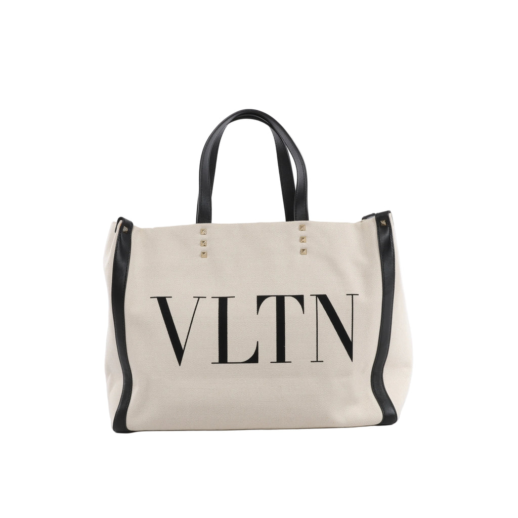 Beige Valentino Garavani VLTN canvas tote bag with black leather handles and trim, oversized black VLTN logo and pyramid stud accents.
