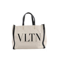 Beige Valentino Garavani VLTN canvas tote bag with black leather handles and trim, oversized black VLTN logo and pyramid stud accents.