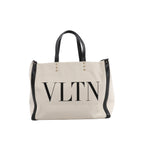 Beige Valentino Garavani VLTN canvas tote bag with black leather handles and trim, oversized black VLTN logo and pyramid stud accents.