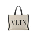 Valentino Garavani VLTN beige canvas tote bag with black leather trim, black top handles, stud details, and large black VLTN logo print on the front.