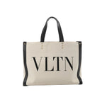 Valentino Garavani VLTN beige canvas tote bag with black leather trim, black top handles, stud details, and large black VLTN logo print on the front.