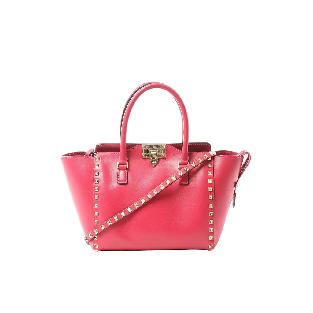 Pink Valentino Garavani Rockstud leather 2-way handbag with top handles, gold-tone stud trim, front turn-lock closure, and studded shoulder strap on white background.