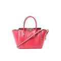 Pink Valentino Garavani Rockstud leather 2-way handbag with top handles, gold-tone stud trim, front turn-lock closure, and studded shoulder strap on white background.