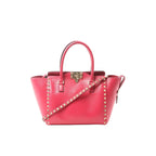 Pink Valentino Garavani Rockstud leather 2-way handbag with top handles, gold-tone stud trim, front turn-lock closure, and studded shoulder strap on white background.